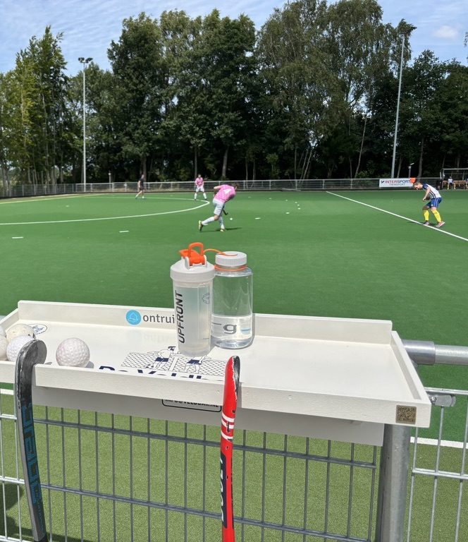 De Veldbar aan het hek langs een hockeyveld, met bidons erop en een wedstrijd op de achtergrond
