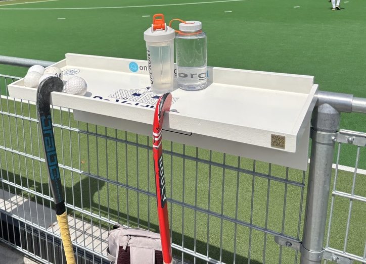 De Veldbar aan het hek met bidons, ballen en hockeysticks erbij, het speelveld erachter